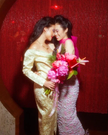 Two women in gowns embrace near a red beaded wall, holding a pink peony bouquet, smiles, and formal wear for an elegant event.