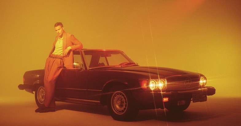 Adult male model leans against a vintage car in warm studio light, wearing a peach suit and yellow shirt against a monochromatic background.