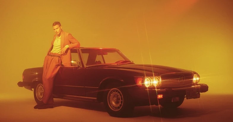 Adult male model leans against a vintage car in warm studio light, wearing a peach suit and yellow shirt against a monochromatic background.