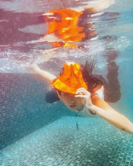 Underwater portrait of a young adult female wearing an orange bucket hat, swimming in a turquoise tiled pool. She is partially submerged.