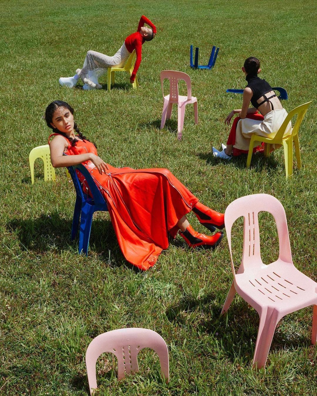 Three women pose in outdoor setting with multiple colorful chairs. Fashion editorial shot with people in colorful outfits on green grass.