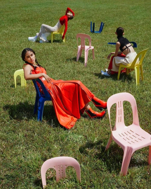 Three women pose in outdoor setting with multiple colorful chairs. Fashion editorial shot with people in colorful outfits on green grass.