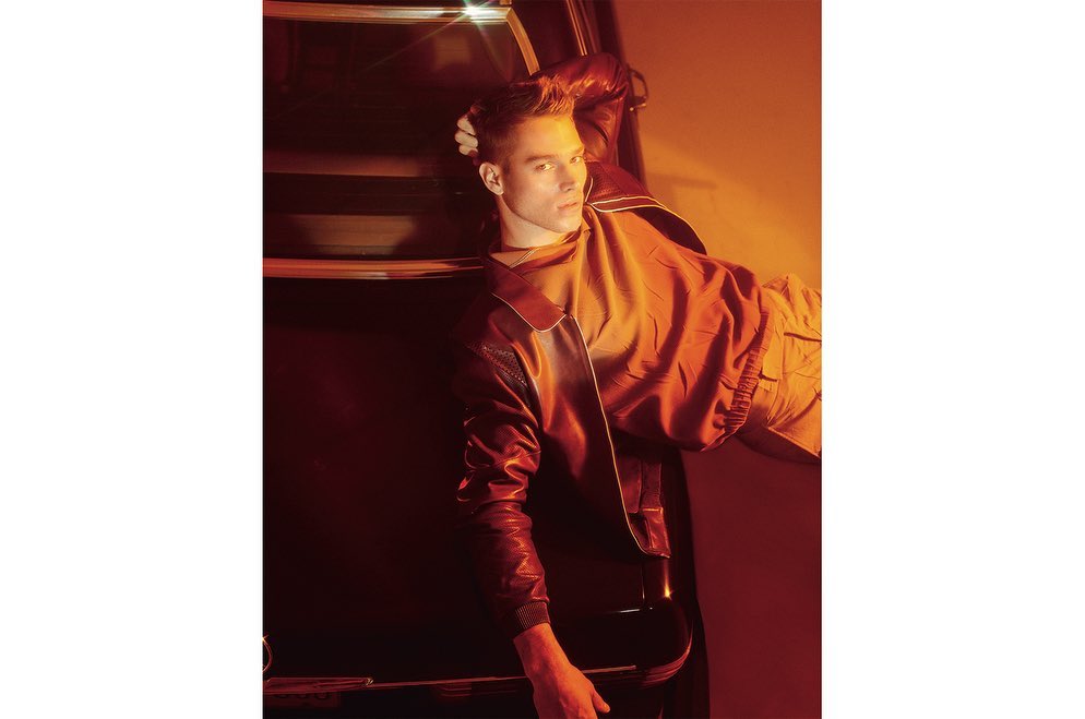 Adult male poses with hand in his hair in studio light wearing a red jacket and khaki pants leaning against a car.