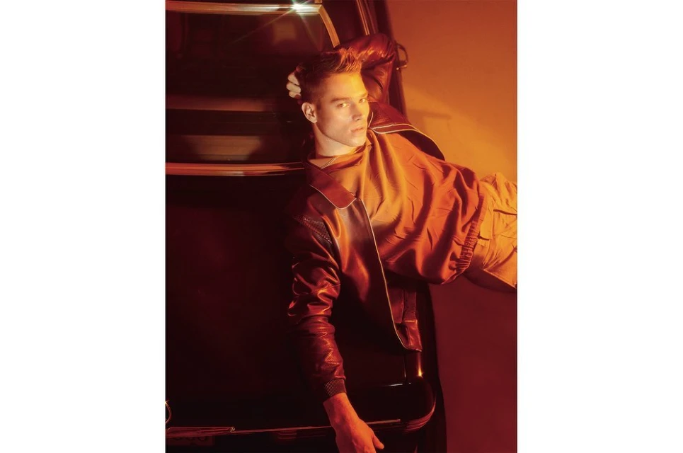 Adult male poses with hand in his hair in studio light wearing a red jacket and khaki pants leaning against a car.