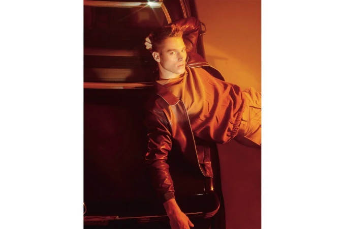 Adult male poses with hand in his hair in studio light wearing a red jacket and khaki pants leaning against a car.
