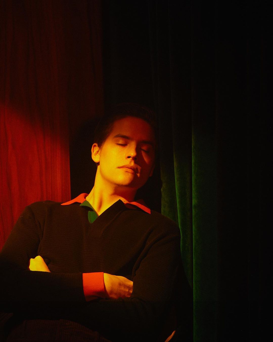 Portrait of a young adult male with closed eyes wearing a black V-neck sweater with orange and green accents against draped fabric.