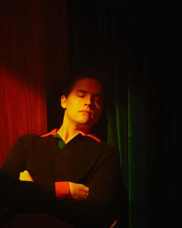 Portrait of a young adult male with closed eyes wearing a black V-neck sweater with orange and green accents against draped fabric.