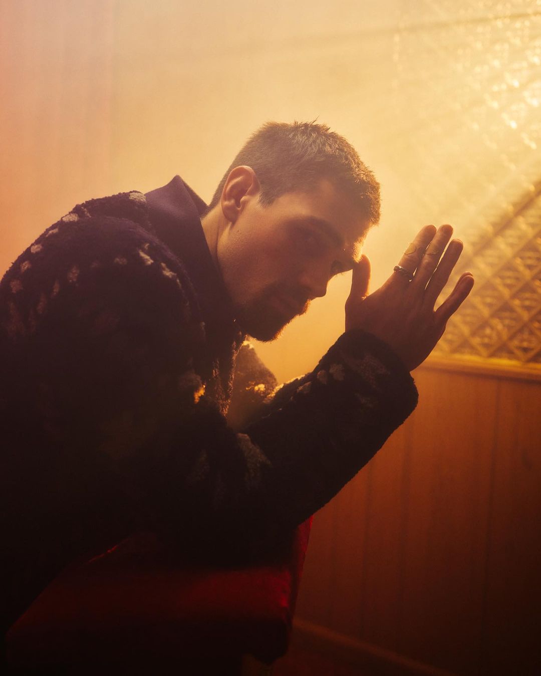 Portrait of an adult man with short brown hair leaning forward with his hand on his forehead indoors in soft, golden light.
