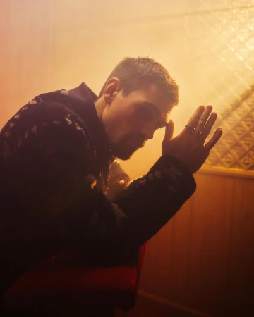 Portrait of an adult man with short brown hair leaning forward with his hand on his forehead indoors in soft, golden light.