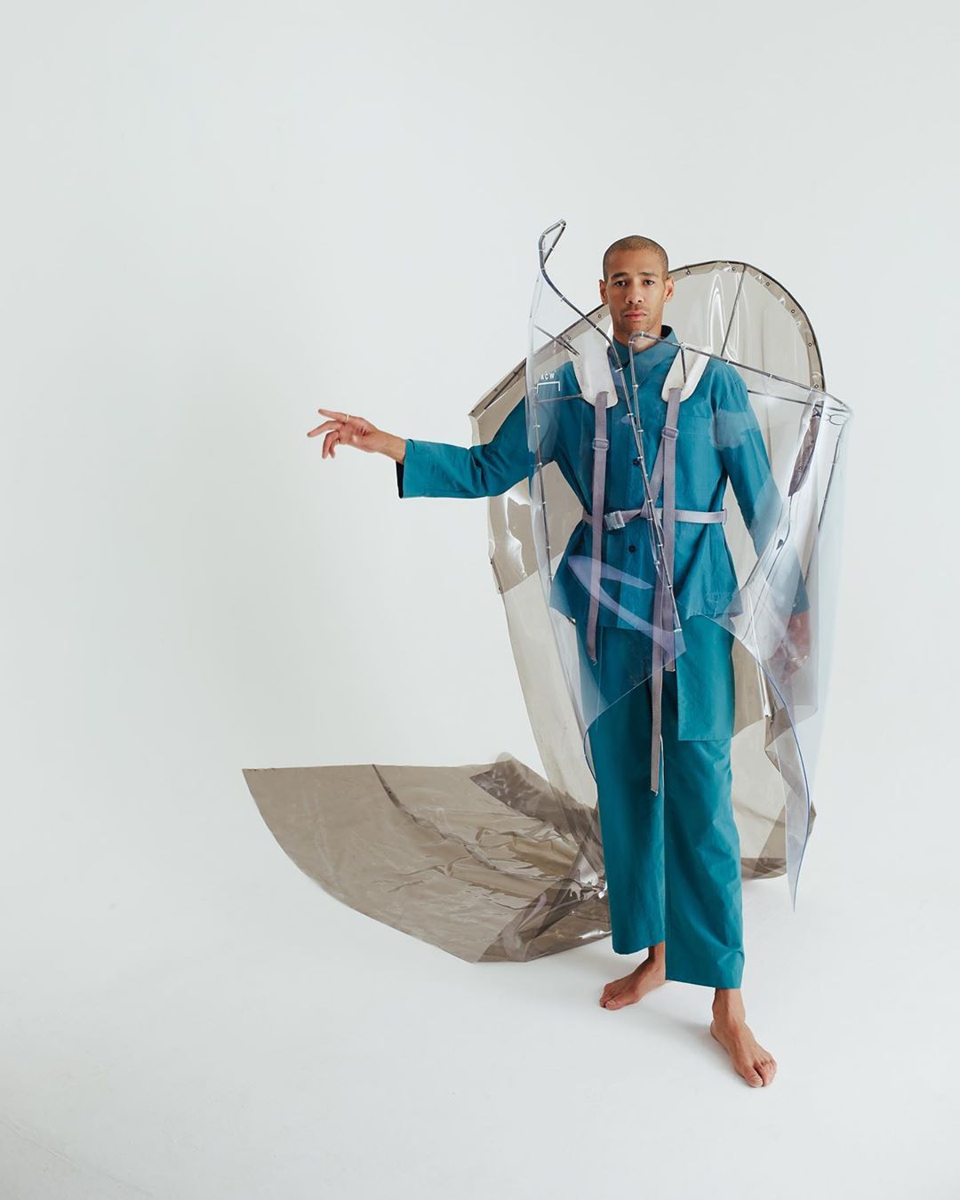 A person wearing an avant-garde transparent garment with blue pants and jacket, standing barefoot in a white studio.