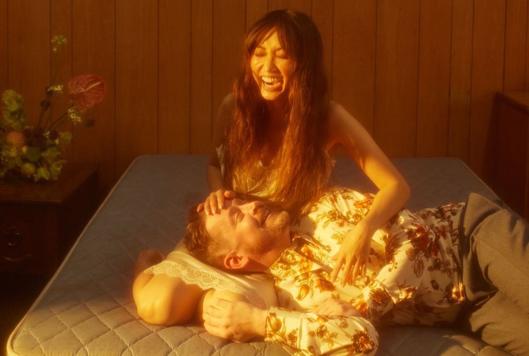 A man lies on a mattress with a woman kneeling next to him, resting her hands on his head. The warm lighting creates a peaceful scene.