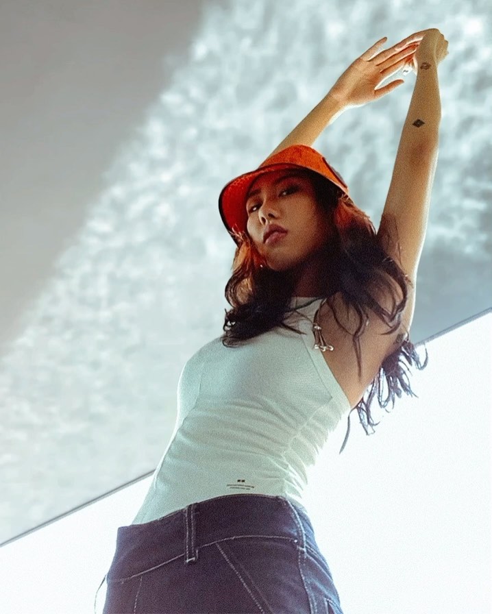 Portrait of a stylish Asian woman in a white top and dark jeans posing with arms raised against a bright neutral background.