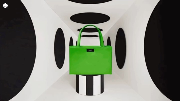 Bright green leather tote bag by Kate Spade, presented on a black-and-white striped pedestal against a geometric backdrop.