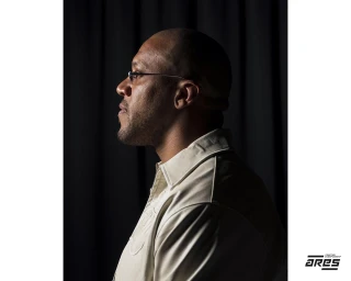Side profile portrait of a man with glasses wearing a cream shirt against a dark curtain backdrop.