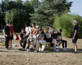 A group of diverse teenagers and young men hanging out around an outdoor concrete table in a sunny park with trees.