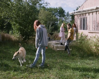 Young woman in a grey hoodie walking a dog in an overgrown grassy garden beside a pale pink house with other people in background.