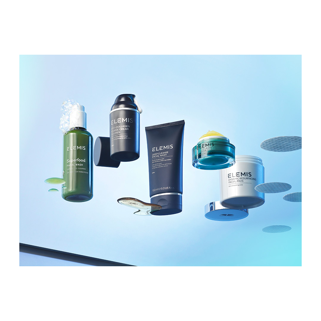 Several Elemis skincare products, including cleanser, cream, facial wash, mask, and pads, are suspended against a gradient blue background.