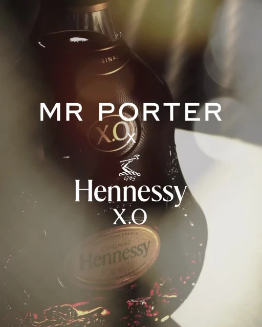 A close-up of a Hennessy X.O cognac bottle, featuring a collaboration with Mr. Porter, set against a blurred, bokeh-lit background.