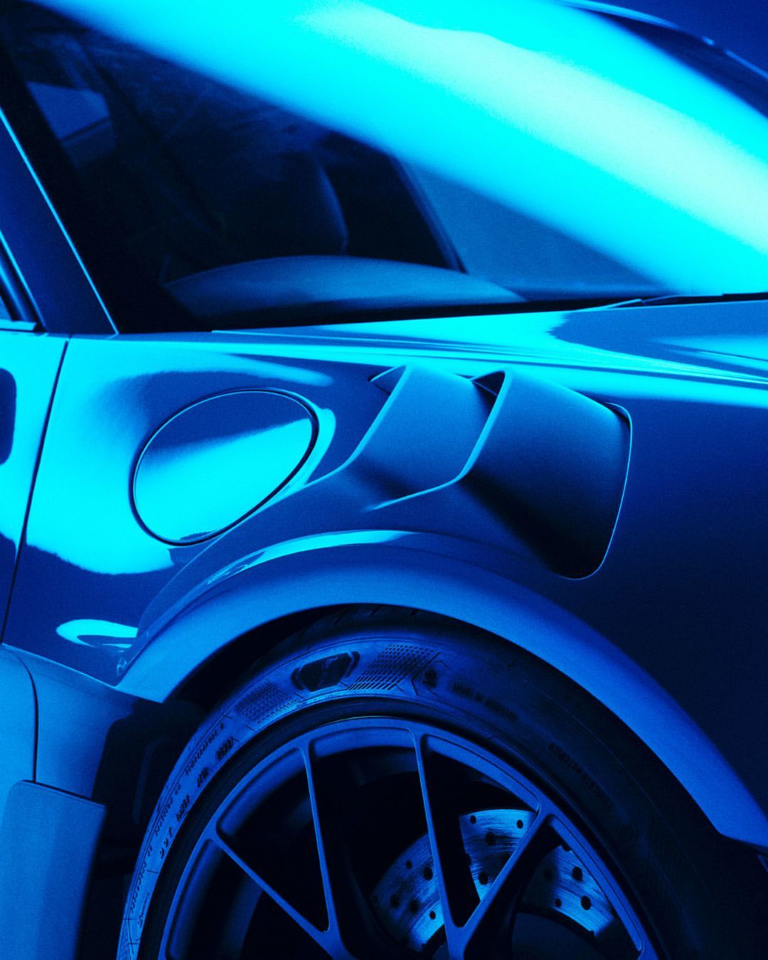 Close-up of a blue luxury car showing the tire, side panel, and ventilation, set in a studio-lit auto industry setting.