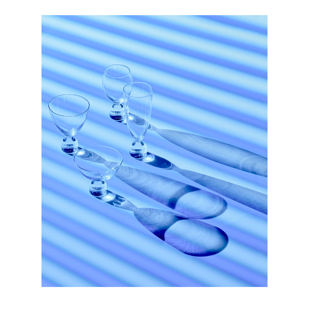 Four empty cocktail glasses stand in a row with distinct shadows on a blue background with horizontal lines in a modern composition.