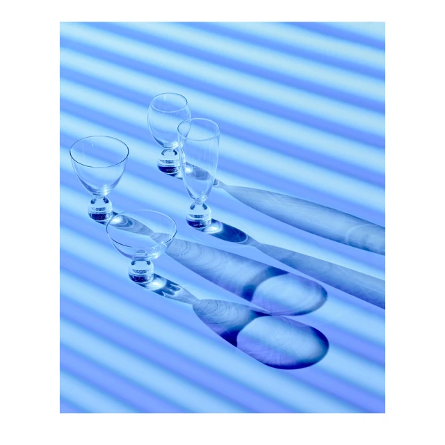 Four empty cocktail glasses stand in a row with distinct shadows on a blue background with horizontal lines in a modern composition.