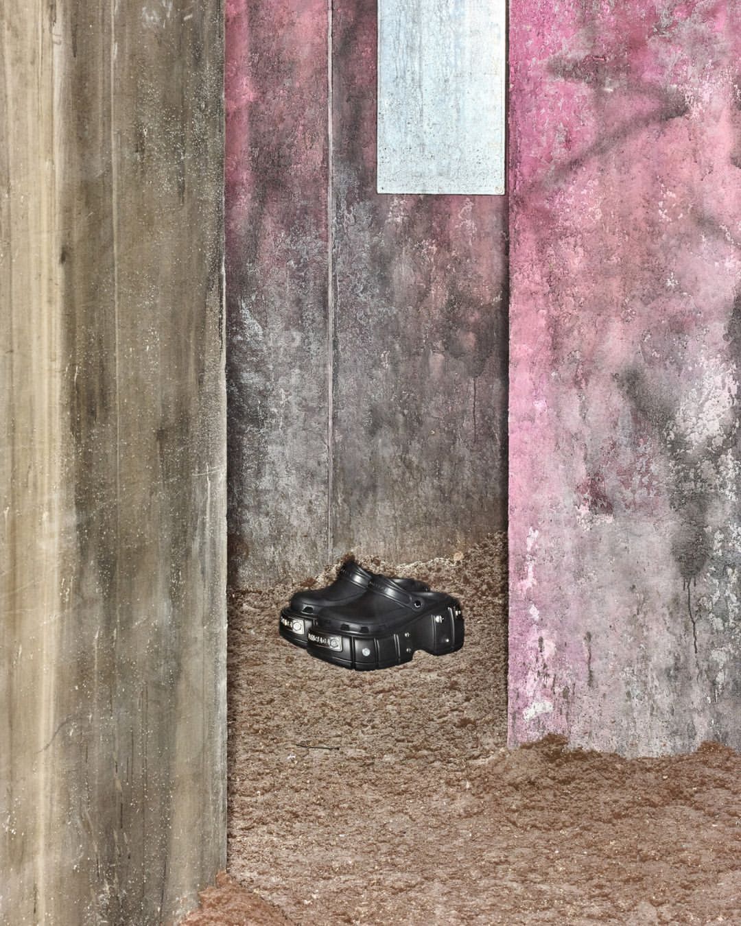 A pair of black platform shoes sit on brown dirt against a textured concrete wall with pink and grey accents, showcasing edgy footwear.
