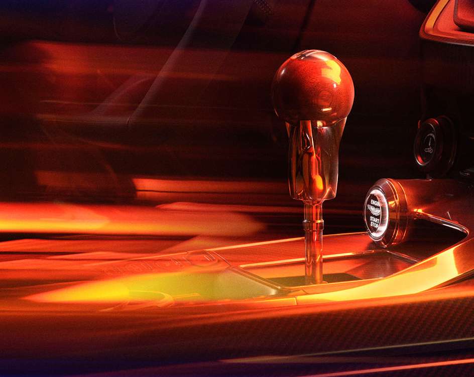 Illuminated interior car detail shot of the gear stick, carbon fiber detailing, and start engine button, with motion-blurred background.