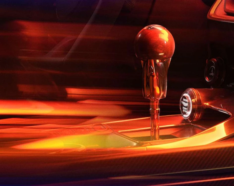 Illuminated interior car detail shot of the gear stick, carbon fiber detailing, and start engine button, with motion-blurred background.