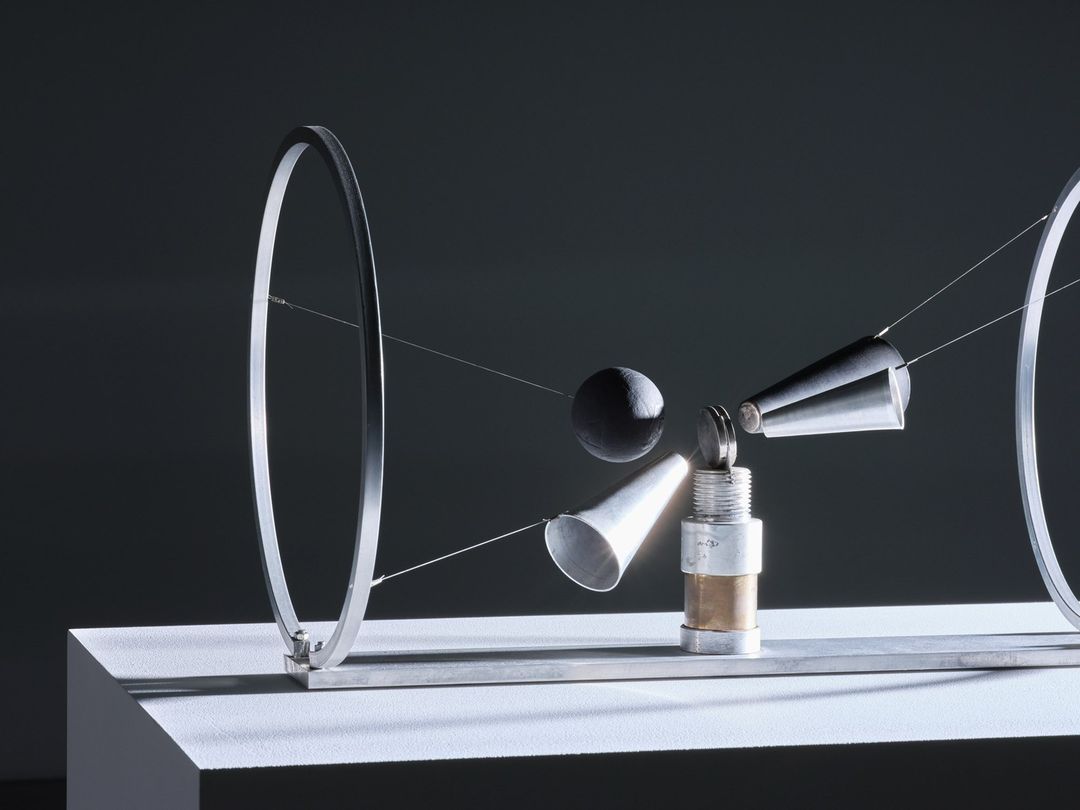 Abstract art piece featuring metal rings, cones, and spheres suspended by wires on a white pedestal against a dark gray background.