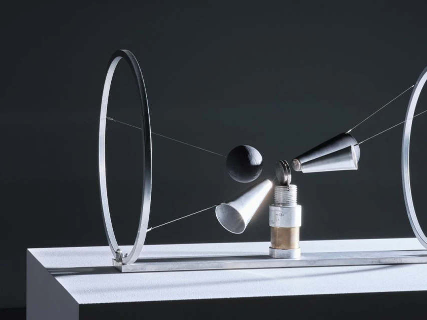 Abstract art piece featuring metal rings, cones, and spheres suspended by wires on a white pedestal against a dark gray background.