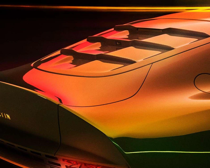 Close-up of the rear of a modern orange and black Aston Martin car with visible taillights, vents, and brand badging on a dark backdrop.