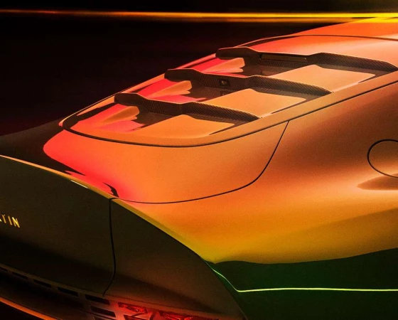 Close-up of the rear of a modern orange and black Aston Martin car with visible taillights, vents, and brand badging on a dark backdrop.