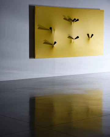 Abstract sculpture art featuring yellow rectangle with multiple black objects creating depth and shadows against a light gray background.