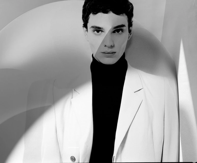 Monochrome portrait of a young man in a turtleneck and suit jacket against a geometric backdrop, showcasing studio lighting.