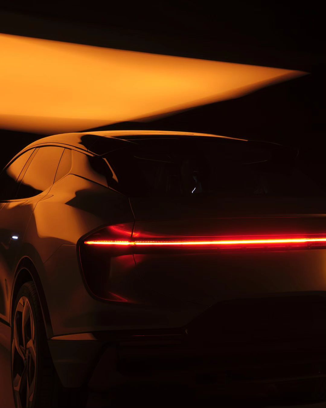 Rear view of a modern silver car with a horizontal red light bar, set against a dark background with soft orange lighting.