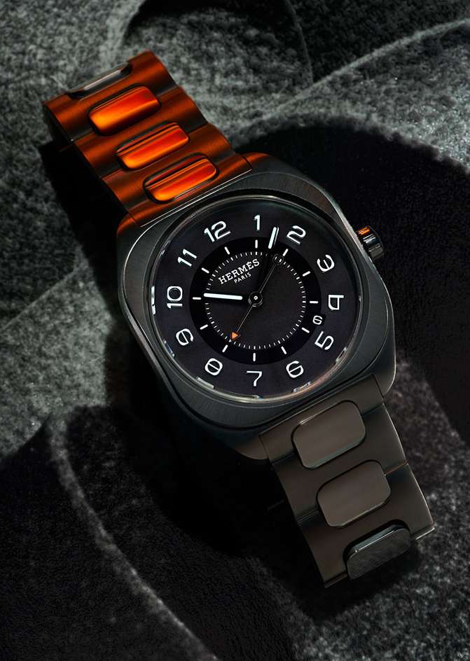 Luxury wristwatch with black dial and unique orange and brown band showcased on a textured grey surface for a high-end fashion appeal.