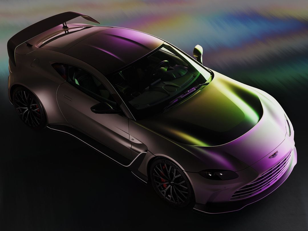 An overhead studio shot showcases a gray Aston Martin Vantage with a rear spoiler, featuring reflections of purple, green, and yellow light.