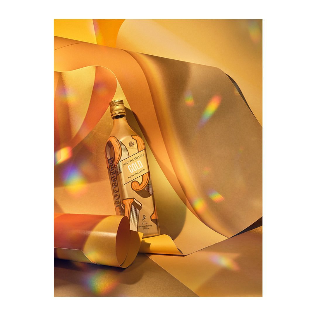 Bottle of Johnnie Walker Gold Label whisky on a golden surface with rainbow prism light and swirling gold paper strips.