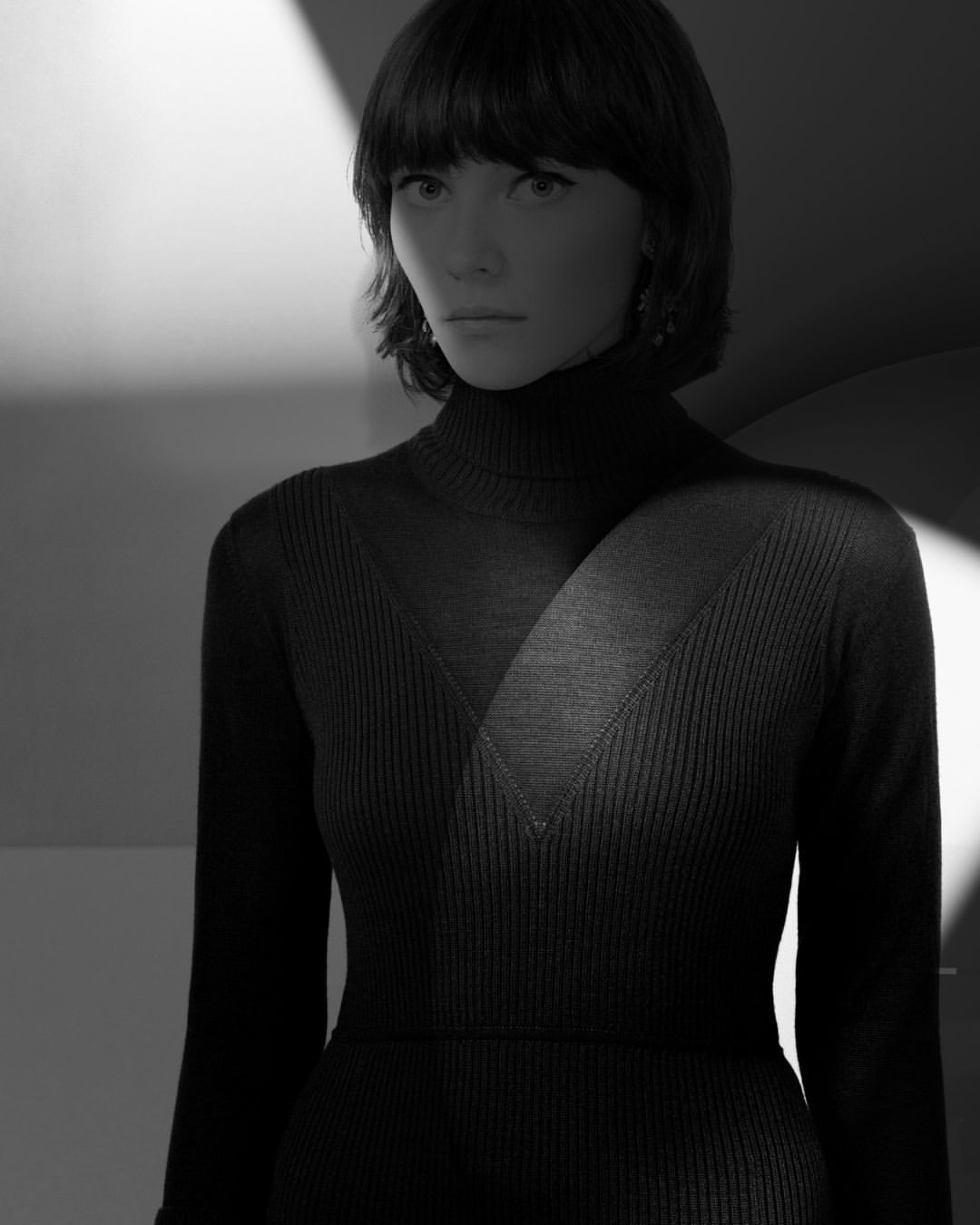 Portrait of an adult woman with bangs wearing a dark turtleneck dress in a photo studio with directed lighting and black and white color grading.