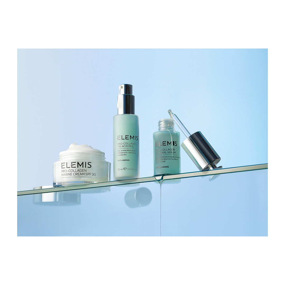 Three ELEMIS skincare product containers on a clear surface against a graduated blue backdrop in a studio shot for beauty advertising.