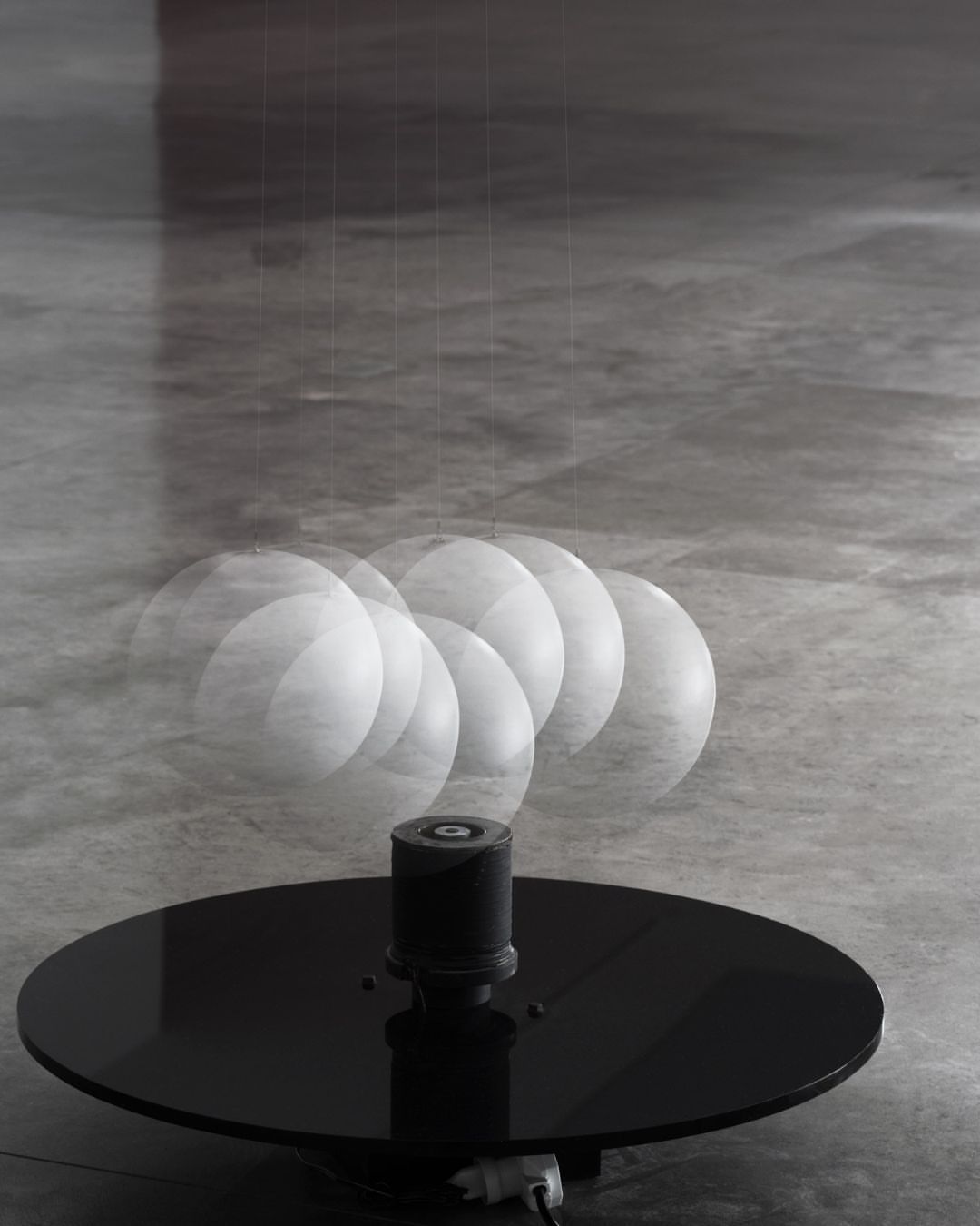 Art installation features a black circular platform with a cylindrical component. A swinging white sphere suspended above creates motion blur.