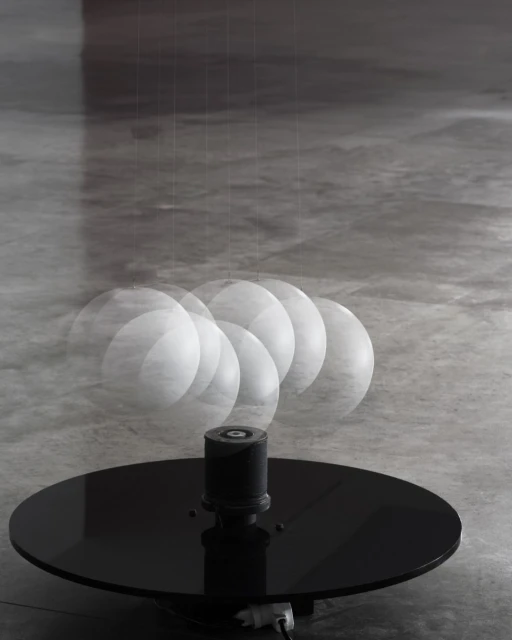 Art installation features a black circular platform with a cylindrical component. A swinging white sphere suspended above creates motion blur.