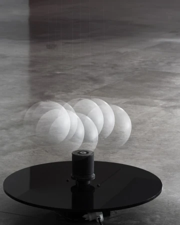 Art installation features a black circular platform with a cylindrical component. A swinging white sphere suspended above creates motion blur.