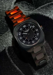 Close up product photograph of a black Hermès wristwatch with a dark textured strap against a grey felt surface.