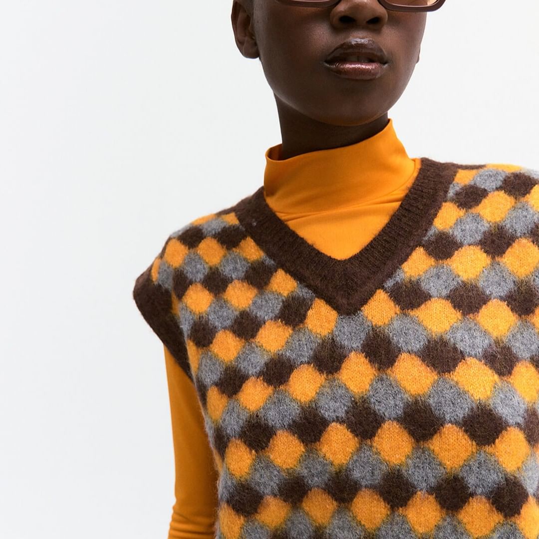An adult African-American woman poses in a photo studio, wearing an orange turtleneck and a brown, orange, and gray checkerboard knit sweater vest.