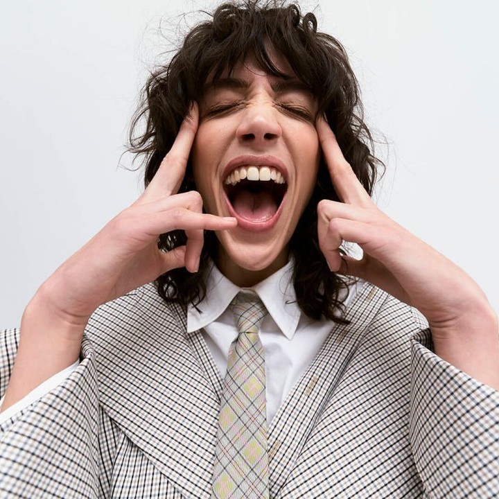 Woman in a checkered blazer and patterned tie poses with her eyes closed and mouth open in front of a plain white background.