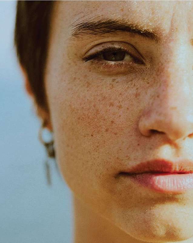 Close-up portrait of a young adult female with freckles, silver earrings, and brown eyes in a natural outdoor setting against a light blue background.
