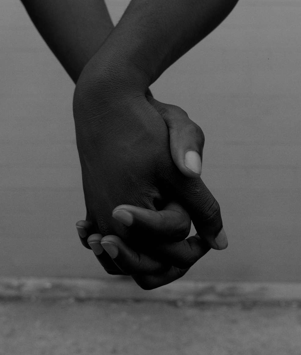Close-up black and white shot of two pairs of human hands, one holding the other, in front of a mottled grey background.