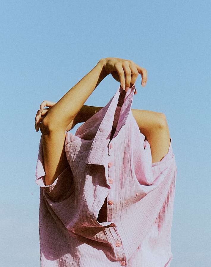 A person stands outdoors against a blue sky, obscured by a pastel purple button-down shirt held overhead, creating a unique fashion pose.