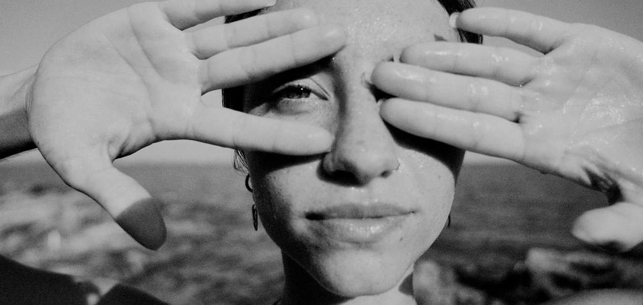Black and white portrait of a young adult female with her hands framing her face against an outdoor background with water and land.